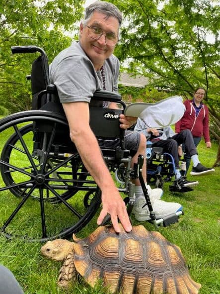 Man with glasses sitting in a wheelchair at Greenburgh Nature Center petting a big turtle.