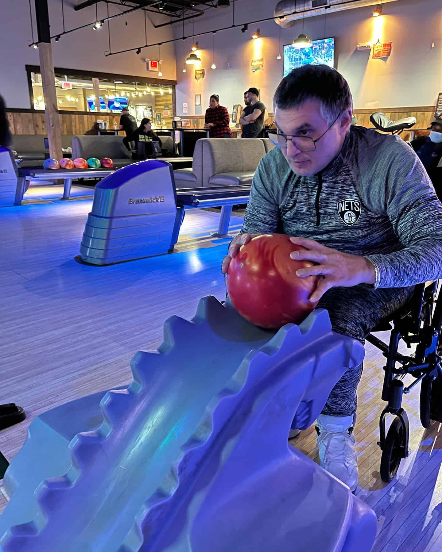 A physically disabled man in a wheelchair getting ready to bowl with an orange bowling ball in a bowling alley.