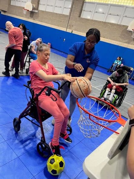 A physically disabled woman sitting in a wheelchair getting ready to dunk a basketball into a hoop.
