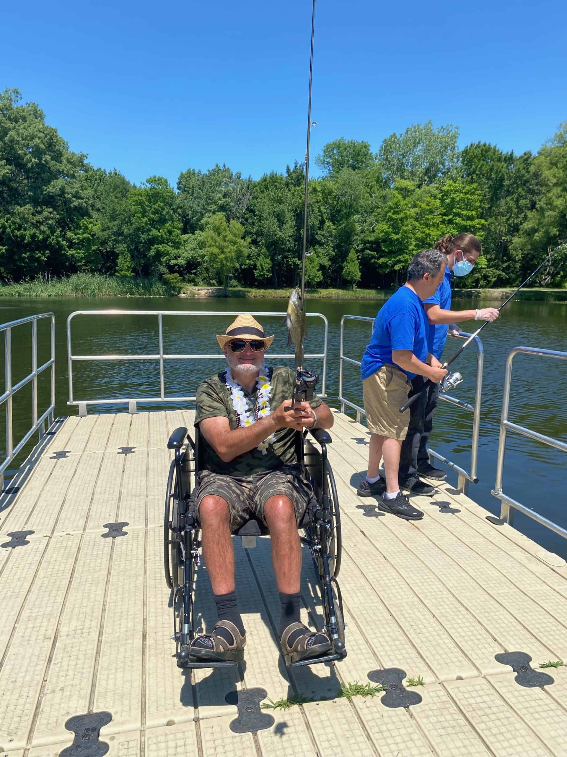 Man wearing dark sunglasses and a straw hat sitting in his wheelchair on a dock holding his fishing pole with a bluegill he caught.